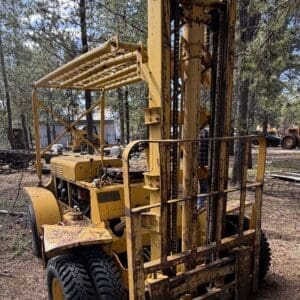IMG_6647 - Montana Auctions by Kevin Hill Old, rusted yellow forklift-like machine in a pine forest, with tall mast, chains, and faded paint awaiting repair.