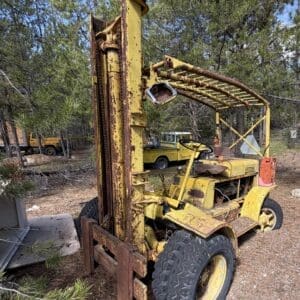 IMG_6651 - Montana Auctions by Kevin Hill Rusty yellow industrial forklift with a tall mast and worn tires, parked among pine trees in a rural lot.
