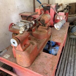 IMG_6658 - Montana Auctions by Kevin Hill Dusty red bench grinder setup on a metal worktable in a cluttered workshop, with hoses and a vise attached.