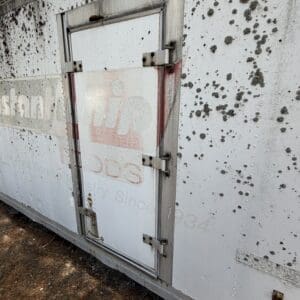 IMG_6663 - Montana Auctions by Kevin Hill Exterior of a weathered white trailer with a vertical rear access door, rusted hinges, and numerous dark spots/speckles on the side, resting on dirt.