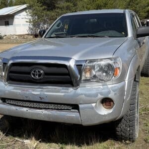 IMG_6670 - Montana Auctions by Kevin Hill Silver Toyota pickup with a damaged front bumper showing peeling paint and scrapes on a dirt lot. The left headlight and grille are visible, and the vehicle has large off-road tires.