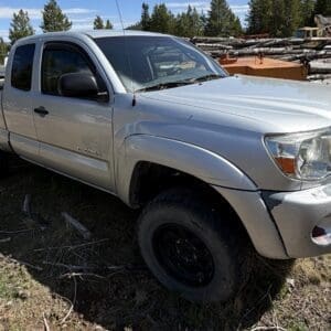 IMG_6672 - Montana Auctions by Kevin Hill Silver Toyota Tacoma pickup with extended cab on a dirt lot, near piles of logs and construction equipment in a rural yard.