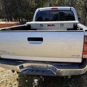 IMG_6673 - Montana Auctions by Kevin Hill Rear view of a silver Toyota Tacoma SR5 pickup with a diamond-plate toolbox in the bed and a hitch, parked on dirt near trees.