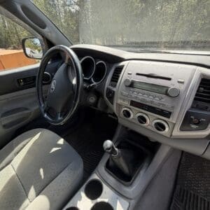 IMG_6676 - Montana Auctions by Kevin Hill Interior of a car with a manual gear shift, central console, and cup holders; sunlit gray upholstery visible from the driver’s seat.