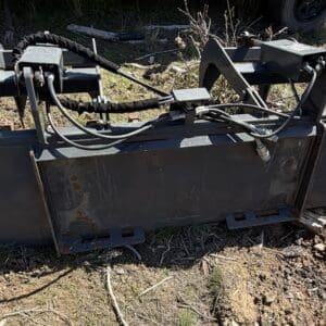 IMG_6686 - Montana Auctions by Kevin Hill Heavy metal skid-steer attachment with hydraulic hoses lying on dirt, likely a bucket/grapple loader attachment awaiting use.