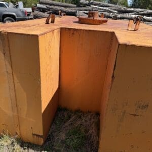 IMG_6689 - Montana Auctions by Kevin Hill Orange, rusted steel industrial container with a large central notch and lifting lugs, outdoors near stacked logs and a pickup truck in the background.