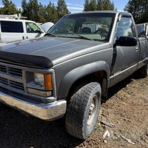IMG_6693 - Montana Auctions by Kevin Hill Gray Chevrolet pickup in a dirt lot with large off-road tires and chrome bumper, surrounded by other vehicles in a salvage yard