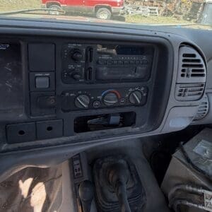 IMG_6697 - Montana Auctions by Kevin Hill Dusty truck cabin dashboard with radio controls and climate dials, visible driver’s legs and boot area, outdoor yard and a red truck seen through the windshield