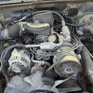 IMG_6700 - Montana Auctions by Kevin Hill Under-hood view of a dusty vehicle engine with belts, hoses, and an alternator visible from the front.