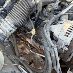 IMG_6701 - Montana Auctions by Kevin Hill Close view of a dusty car engine bay showing a ribbed air intake hose, serpentine belt area, electrical wiring, and an alternator on the right side.