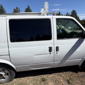 IMG_6705 - Montana Auctions by Kevin Hill White cargo van with ladder rack on top, tinted side windows, parked on gravel with trees in the background.