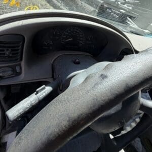 IMG_6707 - Montana Auctions by Kevin Hill Driver's-seat view of a dusty steering wheel, dashboard gauges, and left stalks inside a car, with a sunlit windshield ahead.