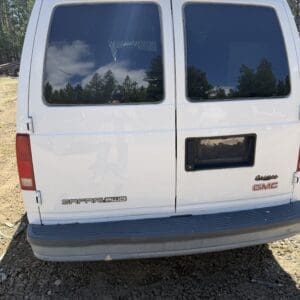 IMG_6710 - Montana Auctions by Kevin Hill White GMC Safari AWD van rear with closed tinted windows, ladder rack on top, parked on dirt with trees in the background; badge reads 'Safari AWD' and 'GMC' on the back doors.