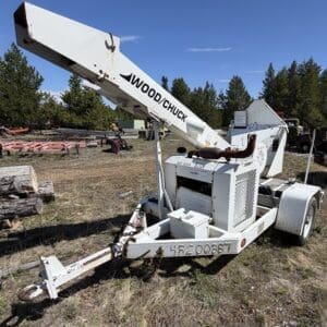 IMG_6711 - Montana Auctions by Kevin Hill Towable white Wood/Chuck log loader crane on a trailer in a rural yard, with logs and pine trees in the background.