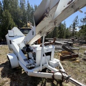 IMG_6714 - Montana Auctions by Kevin Hill White industrial wood chipper on a trailer with a raised feeding chute, parked on a grassy lot with stacked timber nearby.
