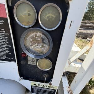 IMG_6715 - Montana Auctions by Kevin Hill Industrial machine control panel with three round gauges (two cracked glass) and a central RPM dial, mounted on a rusting white frame with a caution label below and a run/idle switch nearby.