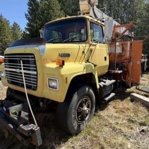 IMG_6716 - Montana Auctions by Kevin Hill Old yellow utility truck with a mounted crane sits in a grassy lot, rusting and weathered.