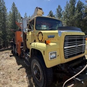 IMG_6720 - Montana Auctions by Kevin Hill Front three-quarter view of a yellow heavy-duty tow truck with a chrome grille and orange equipment on a dry grassy lot, pine trees in the background.