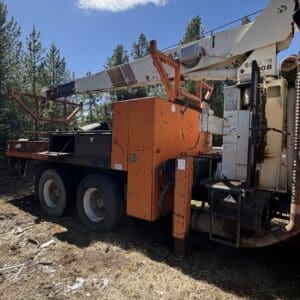 IMG_6721 - Montana Auctions by Kevin Hill Large industrial drilling rig on a truck with an orange control box and white boom, parked on dirt among pine trees under a blue sky.