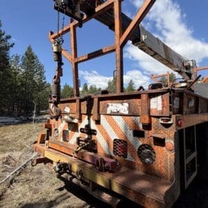 IMG_6722 - Montana Auctions by Kevin Hill Rusted orange utility truck bed with a large crane boom and winch, parked on dirt with trees in the background.