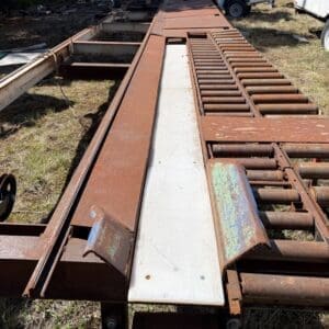 IMG_6735 - Montana Auctions by Kevin Hill Rusty metal roller conveyor frame outdoors, showing a row of rusted cylindrical rollers on the right and a white central panel.
