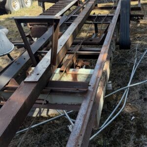 IMG_6740 - Montana Auctions by Kevin Hill Rusty metal trailer frame with long rails and beams lying on dry grass in an outdoor junkyard, with a wheel and cables nearby.