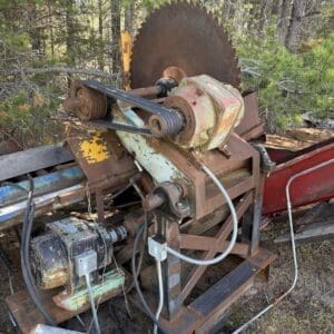 IMG_6741 - Montana Auctions by Kevin Hill Rusty vintage sawmill machine with a large circular blade and belt-driven pulleys, outdoors among trees; motor and wiring visible.