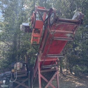 IMG_6742 - Montana Auctions by Kevin Hill Red inclined belt conveyor with metal rollers and gears, mounted on a red frame outdoors among trees.