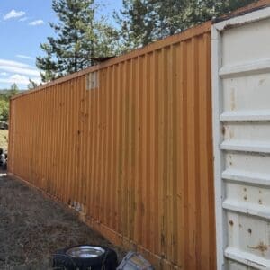 IMG_6745 - Montana Auctions by Kevin Hill Orange corrugated shipping container wall running along a yard, with a white container end on the right; a spare tire and scattered debris sit on dry grass in the foreground.