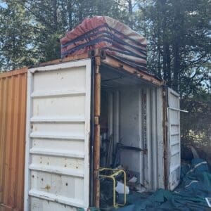 IMG_6746 - Montana Auctions by Kevin Hill Open white shipping container with rusted edges, cluttered interior, and a yellow hand truck inside, outdoors among trees.