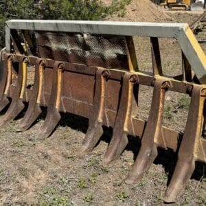 IMG_6747 - Montana Auctions by Kevin Hill Rusty steel rake attachment with multiple curved teeth on a yellow metal frame, resting on dry dirt at a construction site