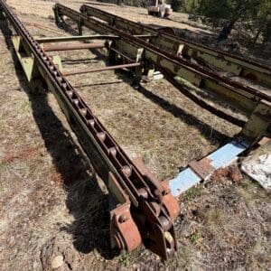 IMG_6753 - Montana Auctions by Kevin Hill Rusted agricultural conveyor system on dry grass with a forest backdrop and distant heavy equipment.