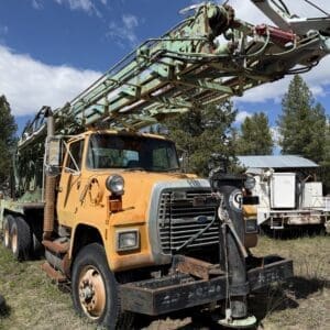 IMG_6754 - Montana Auctions by Kevin Hill Old orange drilling truck with a large green derrick extending overhead on a grassy field under a blue sky, showing rust and wear.