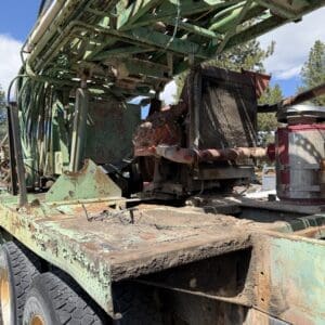 IMG_6755 - Montana Auctions by Kevin Hill Rusty green drilling rig mounted on a flatbed trailer with large tires, outdoors under a blue sky.