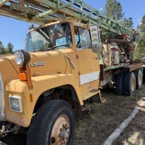 IMG_6759 - Montana Auctions by Kevin Hill Old yellow L9000 truck with a trailer carrying green steel framework, parked on grass under a blue sky.