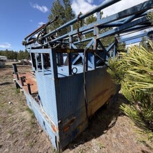 IMG_6765 - Montana Auctions by Kevin Hill Blue, diamond-plate trailer with rust and a long boom/ladder structure, parked in a dry, grassy yard with pine trees in the background.