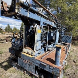 IMG_6766 - Montana Auctions by Kevin Hill Rusty blue drilling rig mounted on a truck in a grassy, outdoor setting with trees in the background