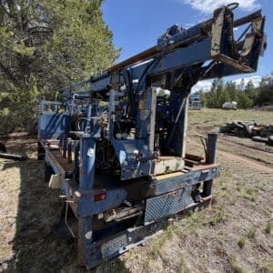 IMG_6767 - Montana Auctions by Kevin Hill Rusted blue industrial drilling rig on a trailer in a grassy clearing, with gauges, hoses, and control levers visible. Pine trees and distant vehicles in the background.