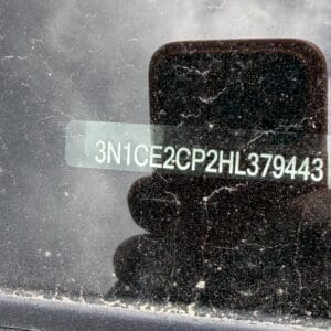 IMG_6780 - Montana Auctions by Kevin Hill VIN sticker on dusty car windshield reads 3N1CE2CP2HL379443, with a phone reflection and dirt on the glass