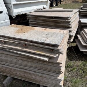 IMG_6782 - Montana Auctions by Kevin Hill Stacks of weathered plywood sheets piled outdoors at a construction site, with trucks in the background.