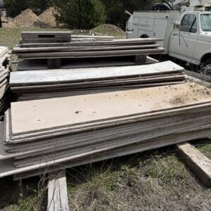 IMG_6783 - Montana Auctions by Kevin Hill Stacks of weathered plywood sheets stacked on wooden pallets at a construction site, with a white utility van and dirt piles in the background.