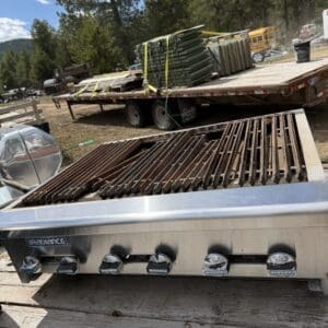 IMG_6784 - Montana Auctions by Kevin Hill Stainless gas grill with heavily used grates on a flatbed trailer in an outdoor yard, with stacked metal rails behind and trees in the background.