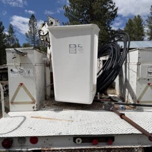 IMG_6788 - Montana Auctions by Kevin Hill Large white electrical cabinet on a flatbed truck with bundled black cables and rusted metal surface, outdoor industrial area.