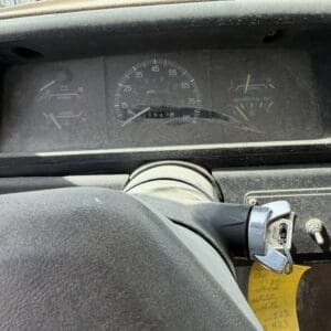 IMG_6792 - Montana Auctions by Kevin Hill Dusty car dashboard with central speedometer, fuel and temperature gauges, steering wheel at bottom, and key in ignition with a yellow tag.