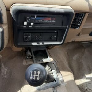 IMG_6794 - Montana Auctions by Kevin Hill Manual gear shift knob for 5-speed (1-3-5 top, 2-4-R bottom) in a beige vehicle interior center console with climate controls and radio behind it.