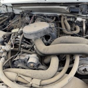 IMG_6796 - Montana Auctions by Kevin Hill Dusty car engine bay with exposed hoses, belts, and the air filter housing under the open hood.