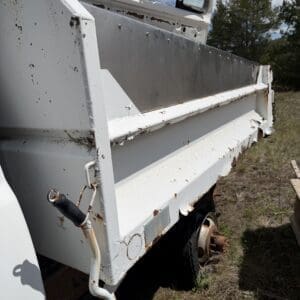 Back of a white, rusting dump truck with peeling paint and exposed wheel hub on dirt ground.”] ,
