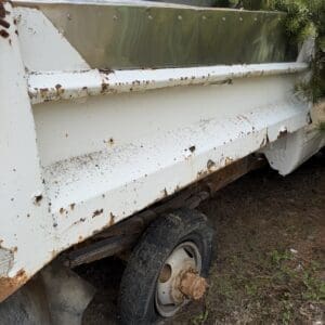 IMG_6801 - Montana Auctions by Kevin Hill Rusted white utility trailer with missing wheel, showing exposed axle and overgrown vegetation nearby