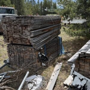 IMG_6803 - Montana Auctions by Kevin Hill Stacks of weathered lumber planks bound with blue straps in a yard, with a dusty vintage truck and scattered metal pipes nearby.