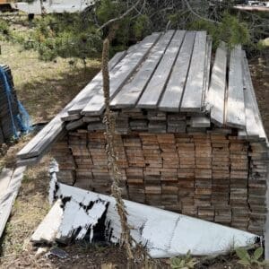 IMG_6804 - Montana Auctions by Kevin Hill Stack of weathered wooden planks neatly piled outdoors under a pine tree, with a bent metal sheet near the base.